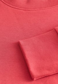 Koraliowy różowy sweter z dzianiny z prążkowanym dekoltem i mankietami, charakteryzujący się miękką teksturą i prostym, bezszwowym designem.