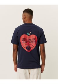 Tamsiai mėlynos spalvos medvilnės marškinėliai, kuriuose pavaizduota didelė raudona širdies grafika su tekstu "LES DEUX LEGACY COURT" ir obuolio motyvas ant nugaros.