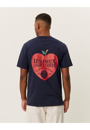Jūras zila kokvilnas T-kreklu ar lielu sarkanu sirds grafiku, uz kuras ir teksts "LES DEUX LEGACY COURT", un ābola motīvu uz aizmugures.