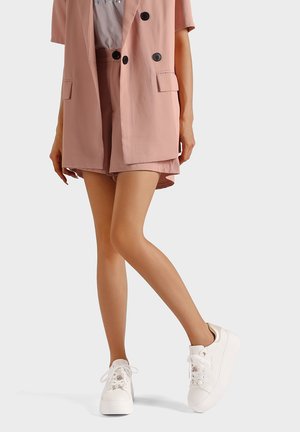 Completo blazer e shorts rosa chiaro in tessuto liscio, abbinati a sneaker bianche con lacci decorativi e suola chunky.