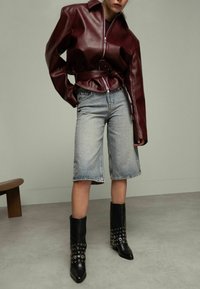 Veste en cuir bordeaux avec une taille ceinturée, associée à un short en denim bleu clair. Des bottines noires à cheville avec des œillets en métal complètent le look.