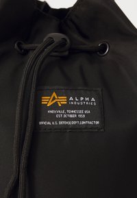 Fekete nylon anyag, húzózsinóros zárással, hímzett címkével, amelyen az "Alpha Industries" felirat és egy narancssárga-fekete embléma látható.