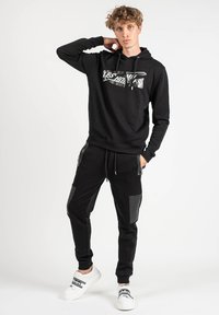 Sudadera negra con estampado gráfico, combinada con joggers negros que tienen bolsillos con cremallera y acentos texturizados en las rodillas, llevada con zapatillas blancas.