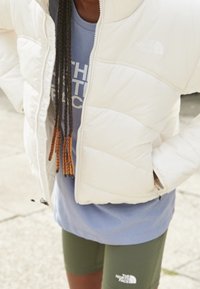 White puffer jacket with horizontal quilted design and a zipper front, worn over a light blue t-shirt. Olive green shorts with branding visible.