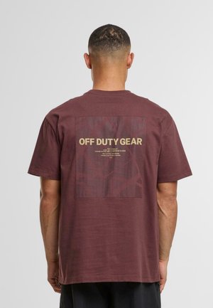 Bărbat îmbrăcat cu un tricou maroniu cu un grafic pătrat și textul "ECHIPAMENT OFF DUTY" pe spate, stând într-un fundal deschis simplu.