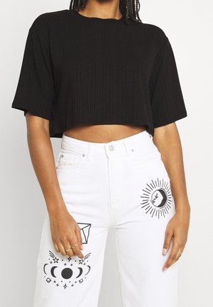 Kobieta ubrana w czarny krótki top i białe spodnie z wysokim stanem z czarnymi motywami niebiańskimi i słonecznymi na udach, stojąca na jednolitym tle.