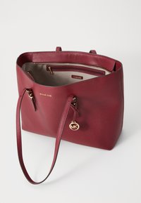 Borsa tote in pelle rossa a forma di ciottoli con due manici robusti, dettagli in metallo dorato e una tasca interna con zip. Presenta un ciondolo con medaglia del logo.