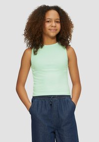 Mintgrünes, geripptes Tanktop mit rundem Ausschnitt, kombiniert mit blauen Denim-Hosen mit Kordelzug. Das Model hat lockige Haare und die Hände in den Taschen.