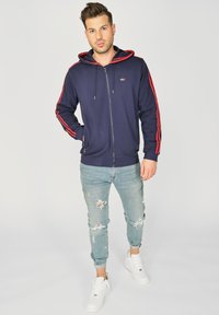 Mørkeblå zip-up hoodie med røde sidelameller, lavet af en bomuldsblanding. Båret med lyseblå slidte jeans og hvide sneakers.