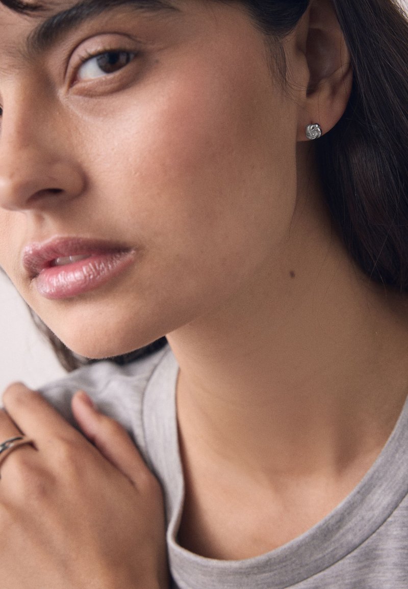 Close-up of a woman wearing a silver knot stud earring and a silver ring, with her hand resting on her shoulder.