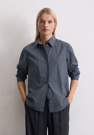 Vrouw met stijlblond haar, gekleed in een zwart overhemd met lichtblauwe geometrische patronen en zwarte broek, staande tegen een witte muur.
