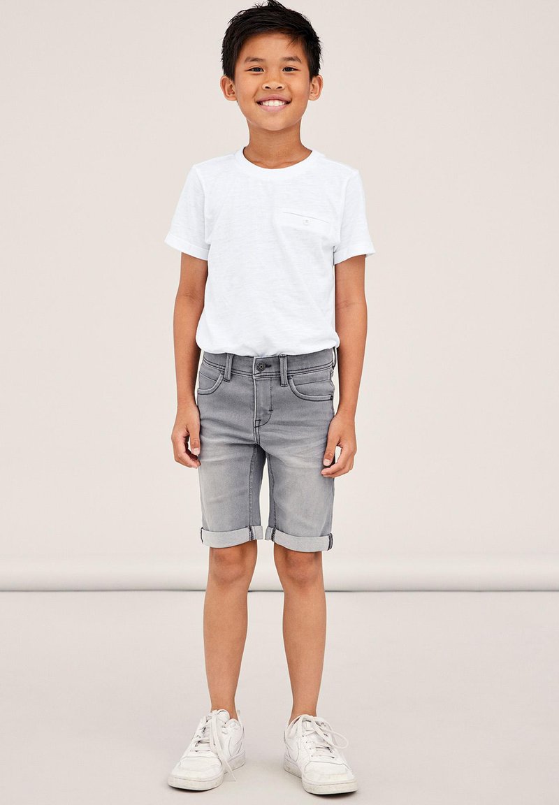 White short-sleeve t-shirt with pocket, paired with light gray rolled-up denim shorts and white sneakers, standing against a neutral background.