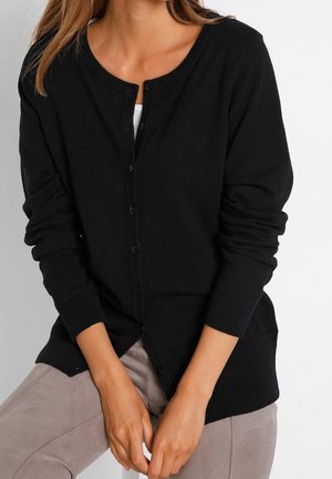 Femme portant un cardigan noir boutonné sur un haut blanc avec un pantalon beige, les mains reposant ensemble devant elle.