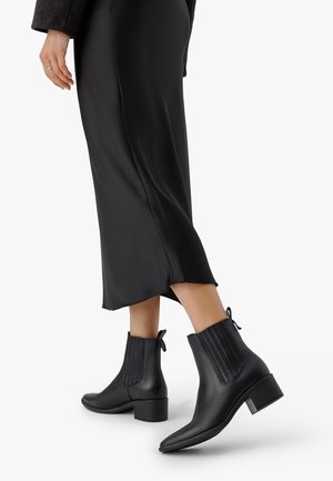 Personne portant des bottines noires à talons bas et une jupe midi noire, faisant un pas en avant sur un fond blanc.