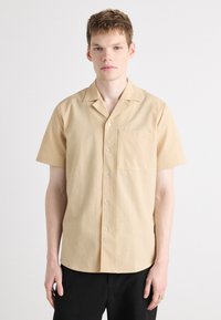 Beige Kurzarmhemd mit strukturiertem Muster, Knopfleiste und einer Brusttasche, kombiniert mit schwarzen Hosen.