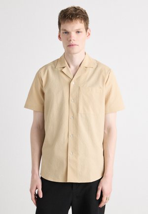 Short-sleeve beige shirt with a textured pattern, button front, and single chest pocket, paired with black pants.