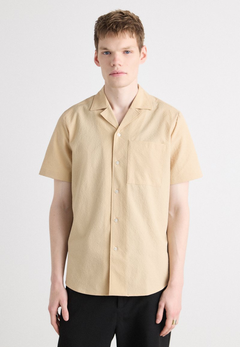 Beige Kurzarmhemd mit strukturiertem Muster, Knopfleiste und einer Brusttasche, kombiniert mit schwarzen Hosen.