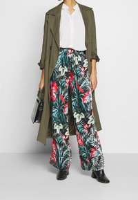 Wijde broek met bloemenprint op een zwarte achtergrond, met levendige rode en groene tropische patronen, gecombineerd met een olijfgroene trenchcoat.
