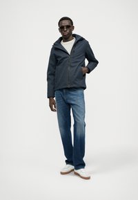 Giacca a cappuccio navy con zip frontale, camicia grigia chiara, jeans blu e sneakers bianche con suole beige, indossata da un modello maschile.