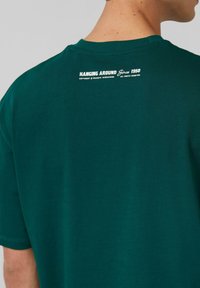Person wearing a dark green T-shirt with white text on the upper back reading "HANGING AROUND Since 1950" and copyright details.