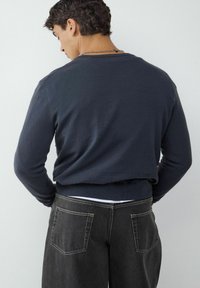 Marineblauw, gebreide trui met lange mouwen en geribbelde zoom; gecombineerd met donkergrijze jeans met dubbele achterzakken en contrasterende stiksels.