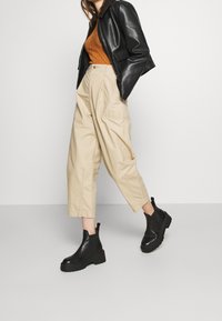 Schwarze Lederjacke, gerippetes oranges Oberteil, beige hochgeschnittene Hose und schwarze Stiefeletten mit klobigen Sohlen. Schlichte, moderne Silhouette.