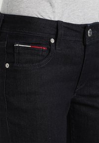 Los jeans de mezclilla oscura presentan un diseño ajustado, un pequeño bolsillo frontal con un acento a rayas en rojo, blanco y azul, y herrajes de botones metálicos.
