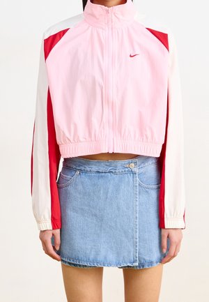 Femme portant une veste courte zippée Nike rose et rouge à manches longues et col montant, associée à une mini-jupe en denim bleu.