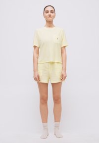 Γυναίκα όρθια που φοράει κοντομάνικο πουκάμισο ανοιχτού κίτρινου χρώματος με ρίγες και κοντάπαντελο σετ, με ανοιχτά μπεζ κάλτσες, μπροστά σε λευκό φόντο.