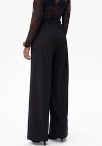 Vrouw met een zwarte wijde high-waist broek, een doorschijnende lange mouwen top met bloemenpatronen en zwarte schoenen met hakken, staand tegen een witte achtergrond.