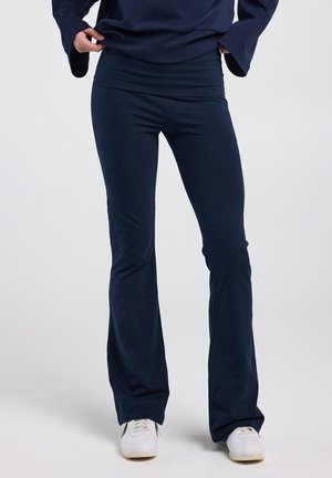 Leggings évasés bleu marine en tissu extensible, dotés d'une large ceinture et d'une texture lisse. Portés avec des baskets blanches.