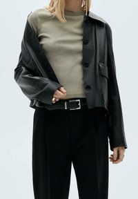 Veste courte en cuir noir avec de gros boutons, associée à une chemise ajustée vert clair et un pantalon noir taille haute. Détail boucle de ceinture argentée.