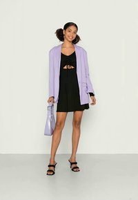 Blazer oversized lilás sobre um vestido mini preto com detalhe em nó, combinado com uma carteira roxa e sandálias pretas com tiras sobre uma superfície clara.