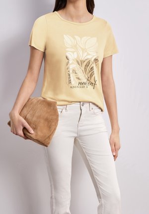 Femme portant un t-shirt graphique floral jaune clair et un pantalon blanc, tenant un sac en daim marron doux dans sa main gauche.