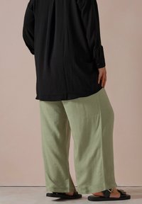 Svart långärmad skjorta, löst sittande ljusgrön texturerad byxor, svarta sandaler med gulddetaljer. Ljus pastellbakgrund.