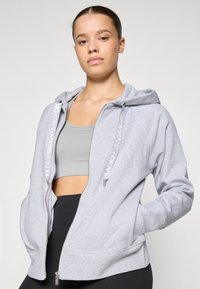 Sweatshirt cinza com fecho, apresentando texto branco nos cordões e bolsos laterais. Textura suave, corte descontraído, combinado com uma t-shirt cinza.