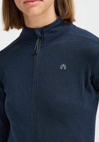 Navy blue zip-up jacket in textured fabric, featuring a high collar, logo on the chest, and dual-tone zipper with black pulls.