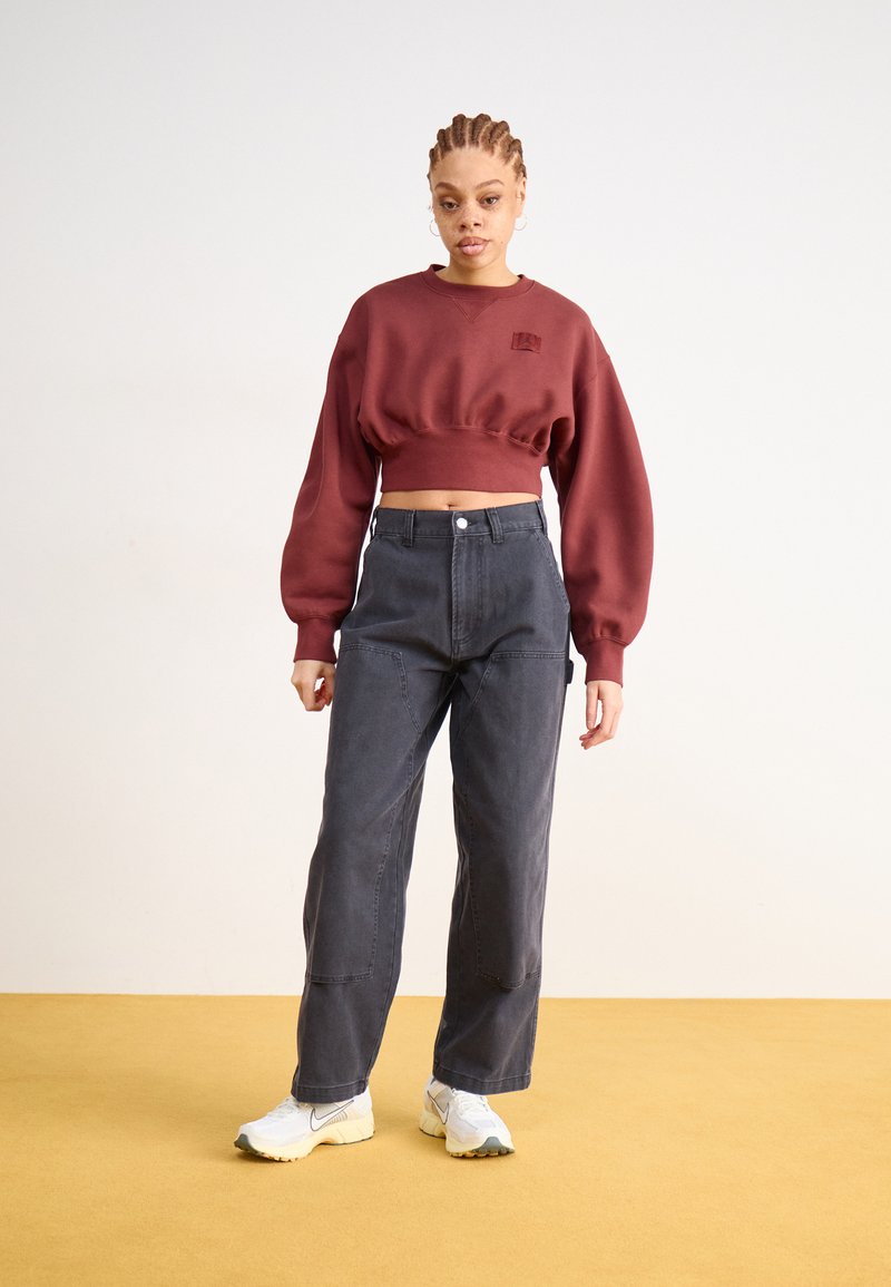 Bordowy skrócony sweatshirt z okrągłym dekoltem, sparowany z ciemnoszarymi spodniami w szerokim kroju, uzupełniony białymi i żółtymi sneakersami na żółtej podłodze.