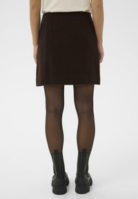 Jupe mini marron en tissu doux, avec une coupe droite. Associée à des collants semi-opaques noirs et des bottes noires à semelle épaisse.