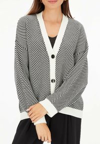 Czarny i biały kardigan w jodełkę z dekoltem w serek, dwiema dużymi czarnymi guzikami i białymi mankietami, wykonany z miękkiej, teksturowanej dzianiny.