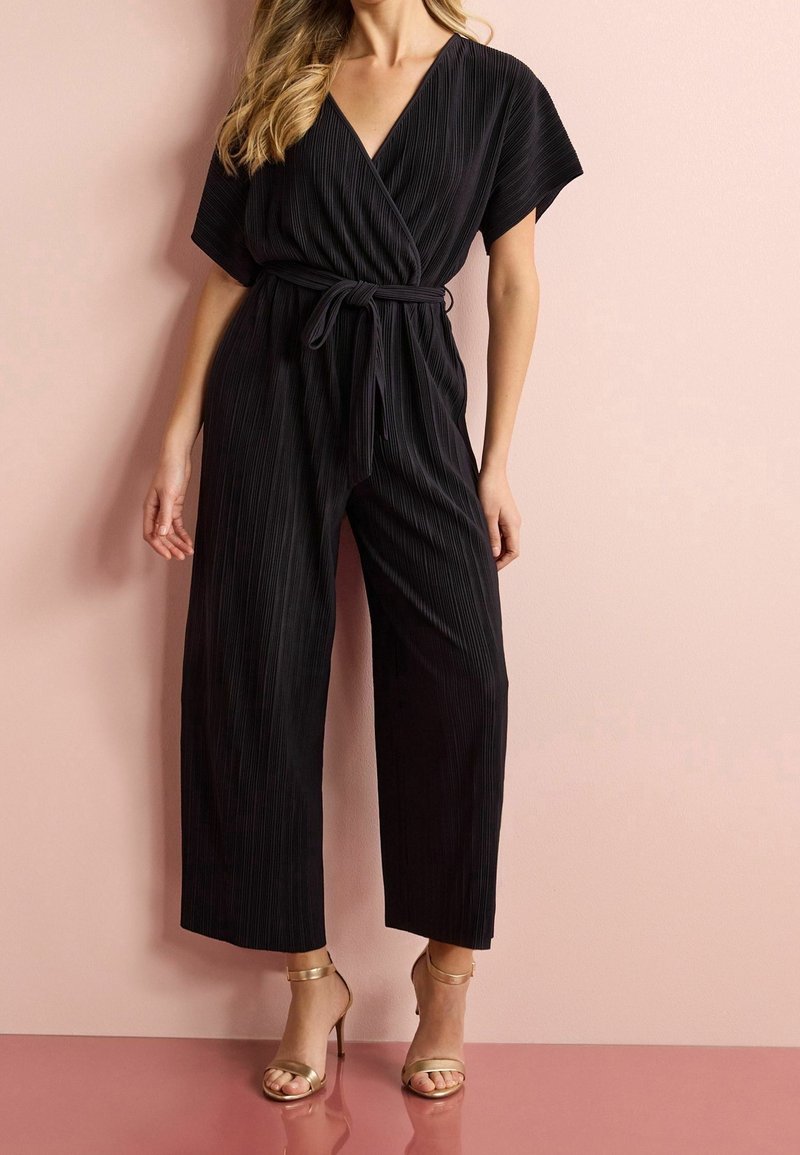 Femme portant une combinaison noire plissée à manches courtes, un col en V, une ceinture à la taille et des sandales dorées à talons hauts, debout contre un mur rose.