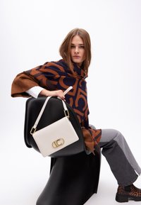 La modella è seduta su una sedia nera, indossando un wrap a righe marrone e blu navy e pantaloni grigi. Tiene in mano una borsa bianca con una chiusura dorata.