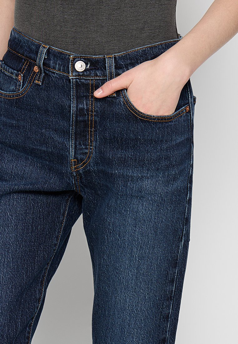Jean en denim bleu foncé à taille haute, avec une poche avant et des coutures orange contrastantes. Il présente un design classique à cinq poches et une fermeture à bouton.
