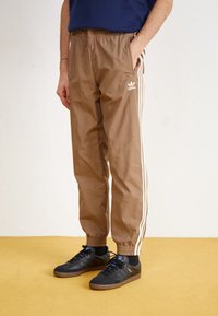 Person som bär bruna Adidas träningsbyxor med vita ränder och svarta Adidas sneakers, stående på ett gult golv mot en vit vägg.