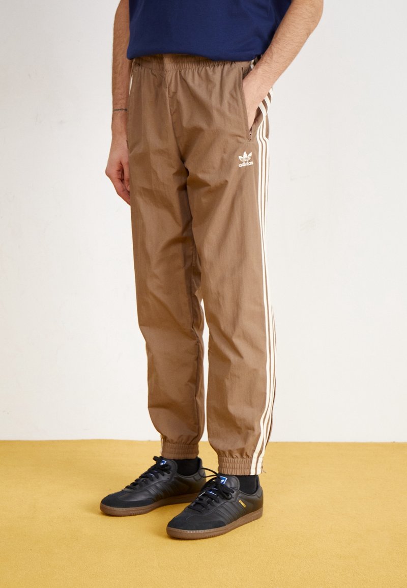 Person som bär bruna Adidas träningsbyxor med vita ränder och svarta Adidas sneakers, stående på ett gult golv mot en vit vägg.