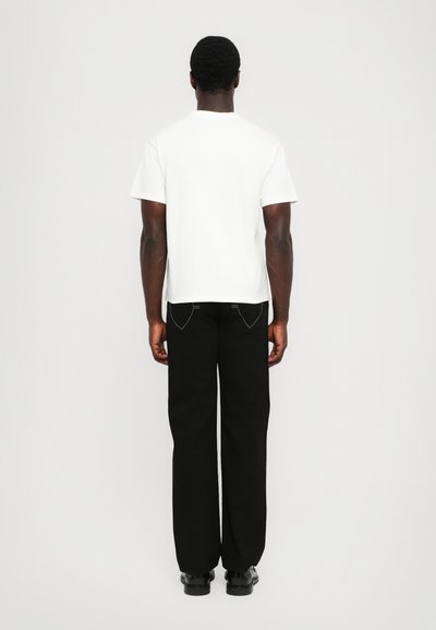 White short-sleeve t-shirt, loose fit, paired with black wide-leg trousers. Visible back pocket stitching detail, smooth fabric texture.