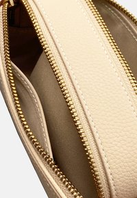 Interno di una borsa in pelle beige testurizzata con doppie cerniere dorate, che mostra un rivestimento in tela liscia e cuciture bianche a contrasto.
