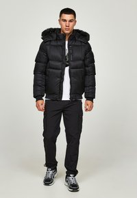 Svart pufferjakke med pelsfôret hette, glidelåslomme på brystet og quiltet mønster. Brukt med svarte cargo-bukser og joggesko.