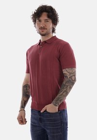 Uomo con capelli ricci che indossa una polo rossa a maniche corte in maglia e jeans blu, mostrando tatuaggi dettagliati su entrambi gli avambracci.