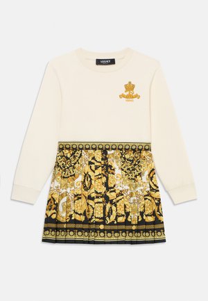 Vestido de manga larga en crema y negro. Presenta patrones florales dorados y una falda plisada. Logotipo bordado en el pecho.